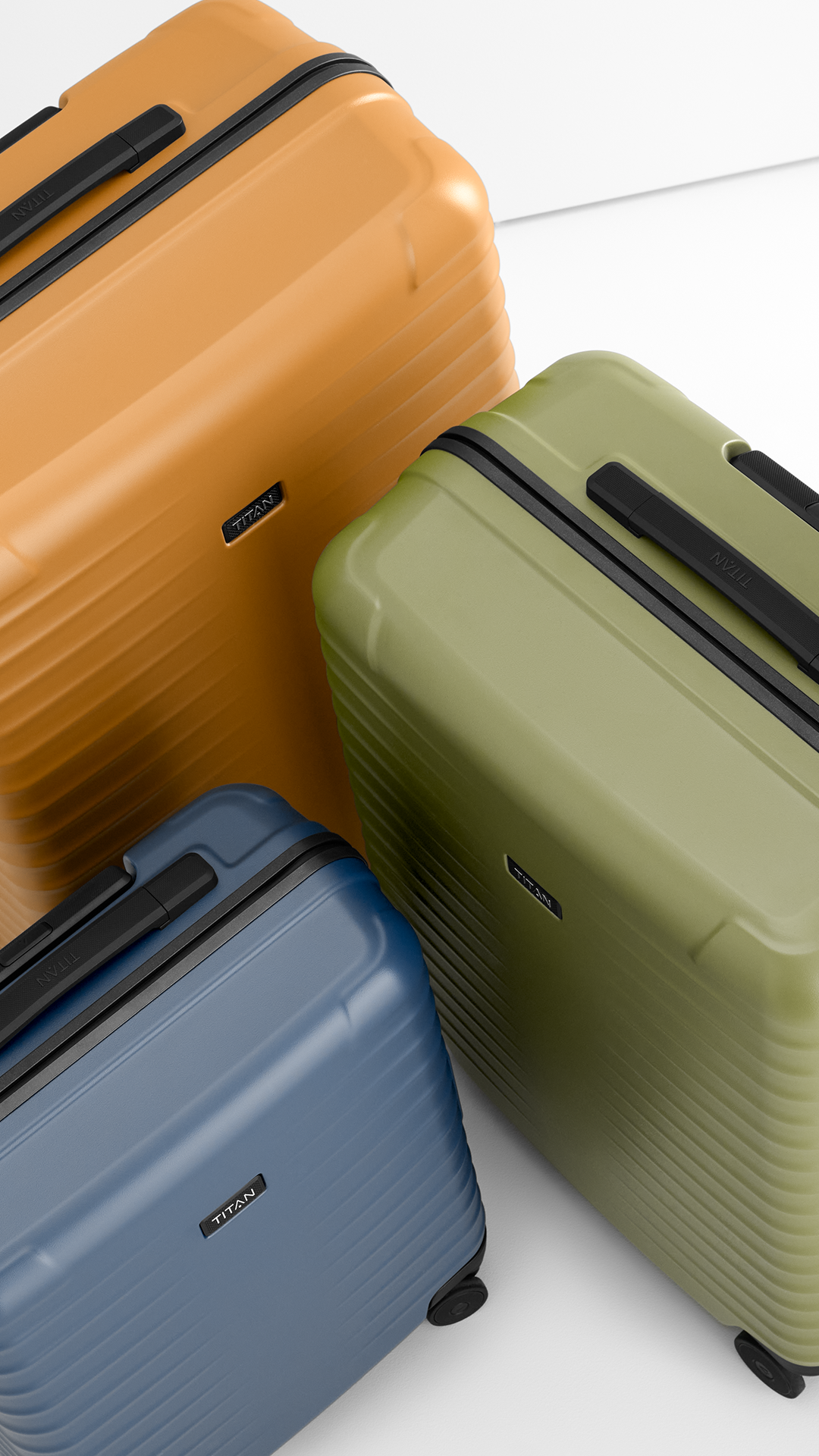 Drei Titan-Koffer in den Farben Orange, Olive und Blau stehen auf einer weißen Oberfläche. Die Koffer haben geriffelte Oberflächen und schwarze Griffe. Das Markenlogo "TITAN" ist dezent auf jedem Koffer angebracht. Die Perspektive ist leicht erhöht.