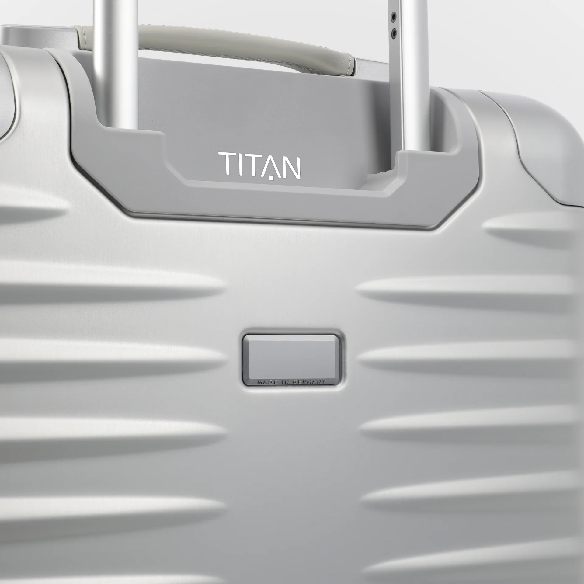 Primer plano de una maleta plateada TITAN. Los detalles incluyen el asa telescópica, el logotipo TITAN y el etiquetado "Made in Germany". La superficie está adornada con líneas horizontales en relieve.