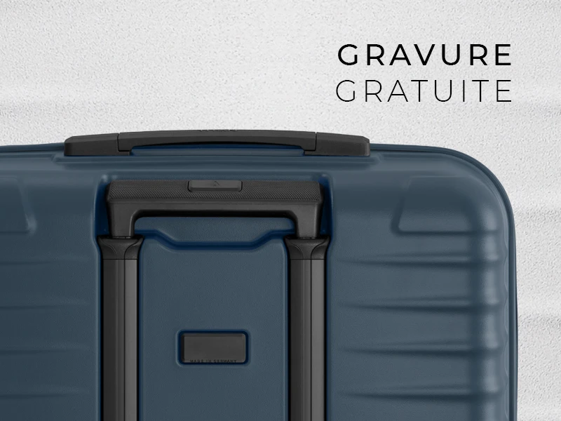 Détail d'une valise de voyage bleue avec poignée et barre d'extension noires. En haut, on peut lire "GRAVURE GRATUITE" en noir. La valise a une surface rainurée et une étiquette avec "MADE IN GERMANY".
