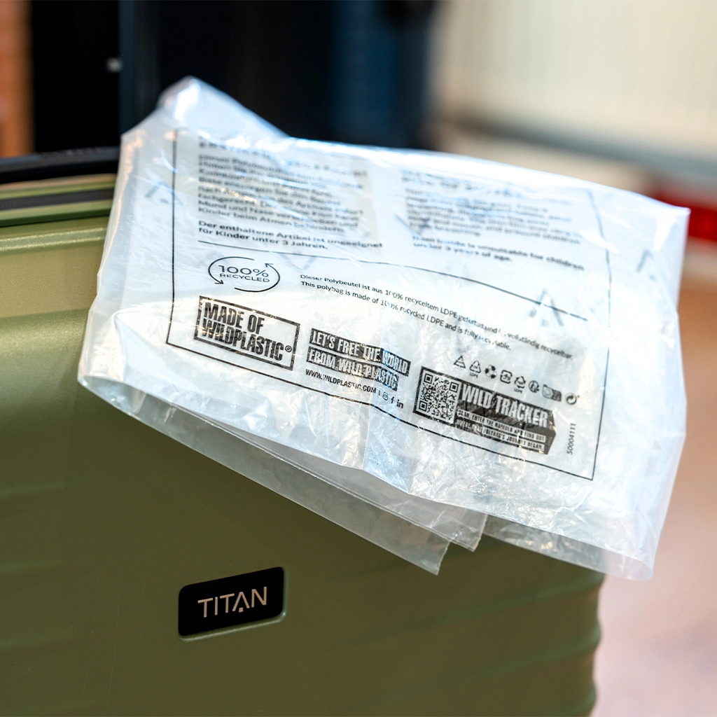 Eine durchsichtige Plastiktüte mit Texten und Logos liegt auf einem olivgrünen Reisekoffer. Auf der Tüte sind unter anderem die Logos "Made of Wildplastic" und "Wild Tracker" zu sehen. Der Koffer hat ein schwarzes Etikett mit dem Schriftzug "TITAN".
