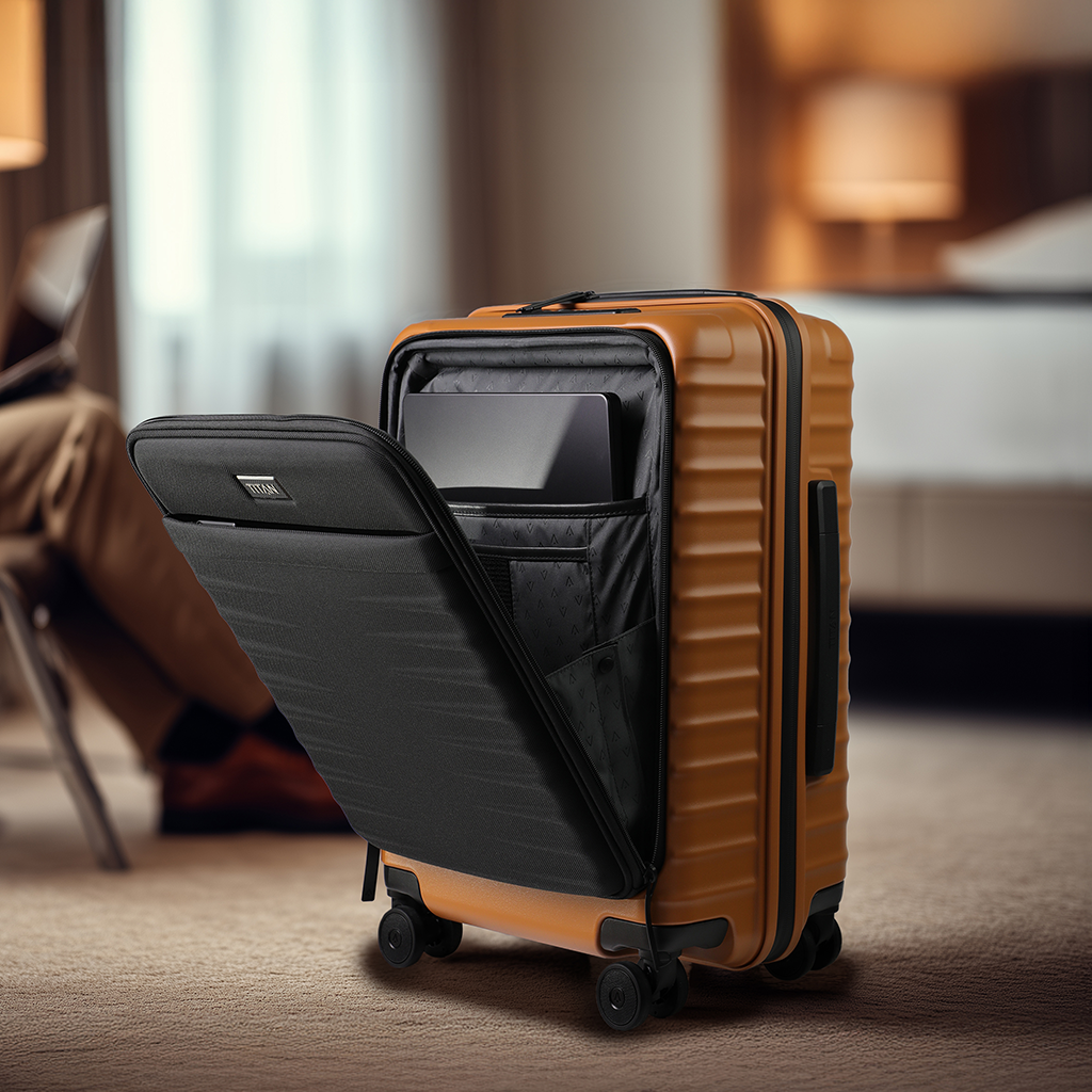 Ein Rollkoffer in Braun mit schwarzen Akzenten steht auf einem Teppich. Die vordere Tasche ist geöffnet und zeigt einen schwarzen Innenraum mit einem Laptop. Im Hintergrund sind verschwommene Möbel und eine Person mit einem Laptop zu sehen.