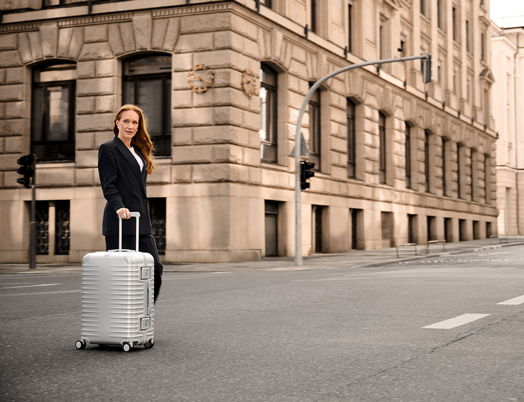 Frau im Business-Outfit zieht einen silbernen Aluminium-Koffer auf einer leeren Straße vor einem historischen Gebäude