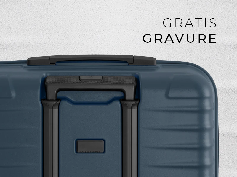 Close-up van een blauwe koffer met zwart handvat en uitschuifbaar telescopisch handvat. Rechtsboven staat de tekst "GRATIS GRAVURE" in zwarte letters. Het oppervlak van de koffer is licht gestructureerd.