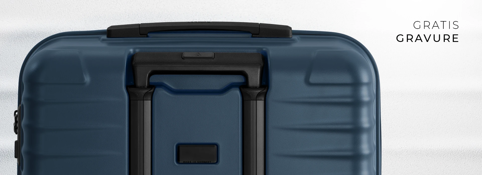 Detailopname van een donkerblauwe koffer tegen een witte achtergrond. De koffer heeft een zwart handvat aan de bovenkant en een uitschuifbaar handvat met zwarte spijlen in het midden. Rechtsboven staat "GRATIS GRAVURE".