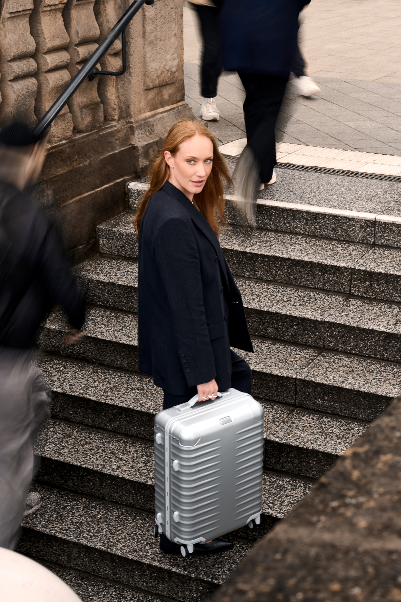Business-Frau trägt einen silbernen Aluminium-Koffer eine steinerne Treppe hinunter, umgeben von Passanten in urbaner Umgebung