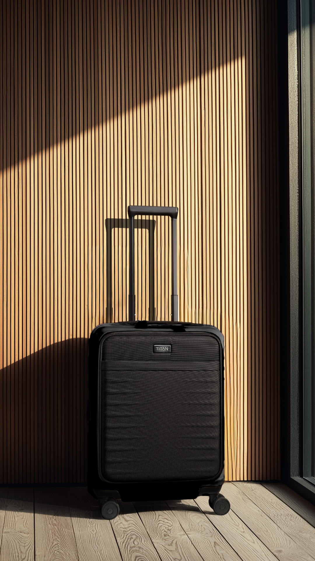 Ein schwarzer Rollkoffer von Titan steht auf einem Holzboden vor einer Wand aus vertikalen Holzpaneelen. Sonnenlicht fällt diagonal auf die Wand und erzeugt ein helles und dunkles Muster. Der Koffer hat zwei ausgezogene, schwarze Teleskopgriffe.